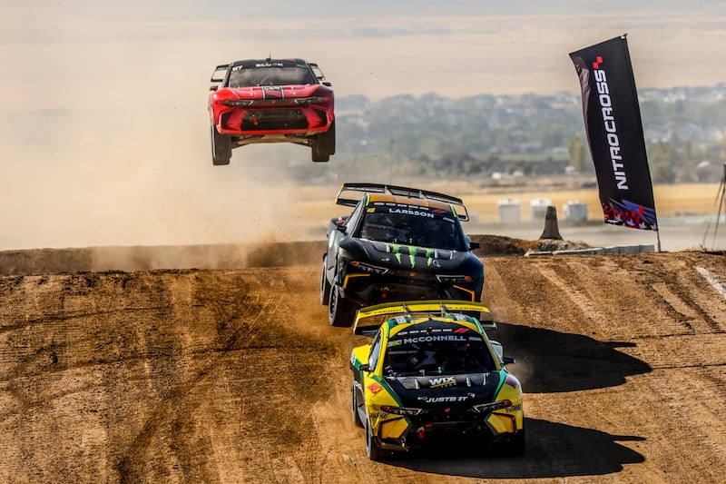 Nitrocross cars racing on dirt track over jump.